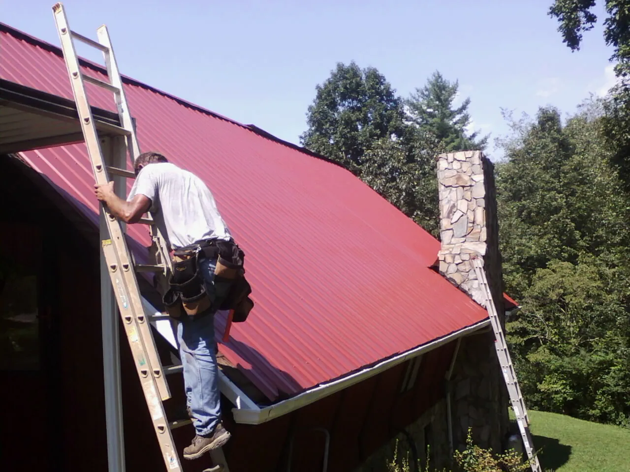 Roofer on a ladder beside a red metal roof and stone chimney — Soffit & Fascia Repair by Roofing Brothers in Clover